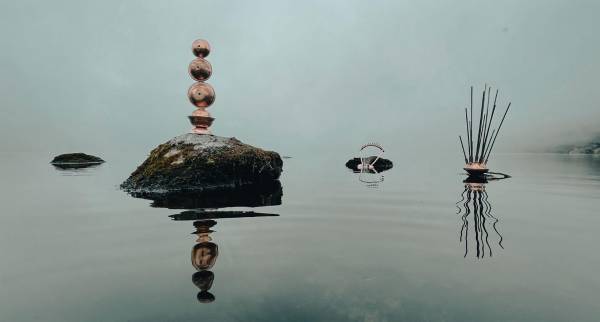 A reflective lake with mossy stones against a great fog. Three brass metal instruments lay on top of the rocks; their reflections ripple in the water.