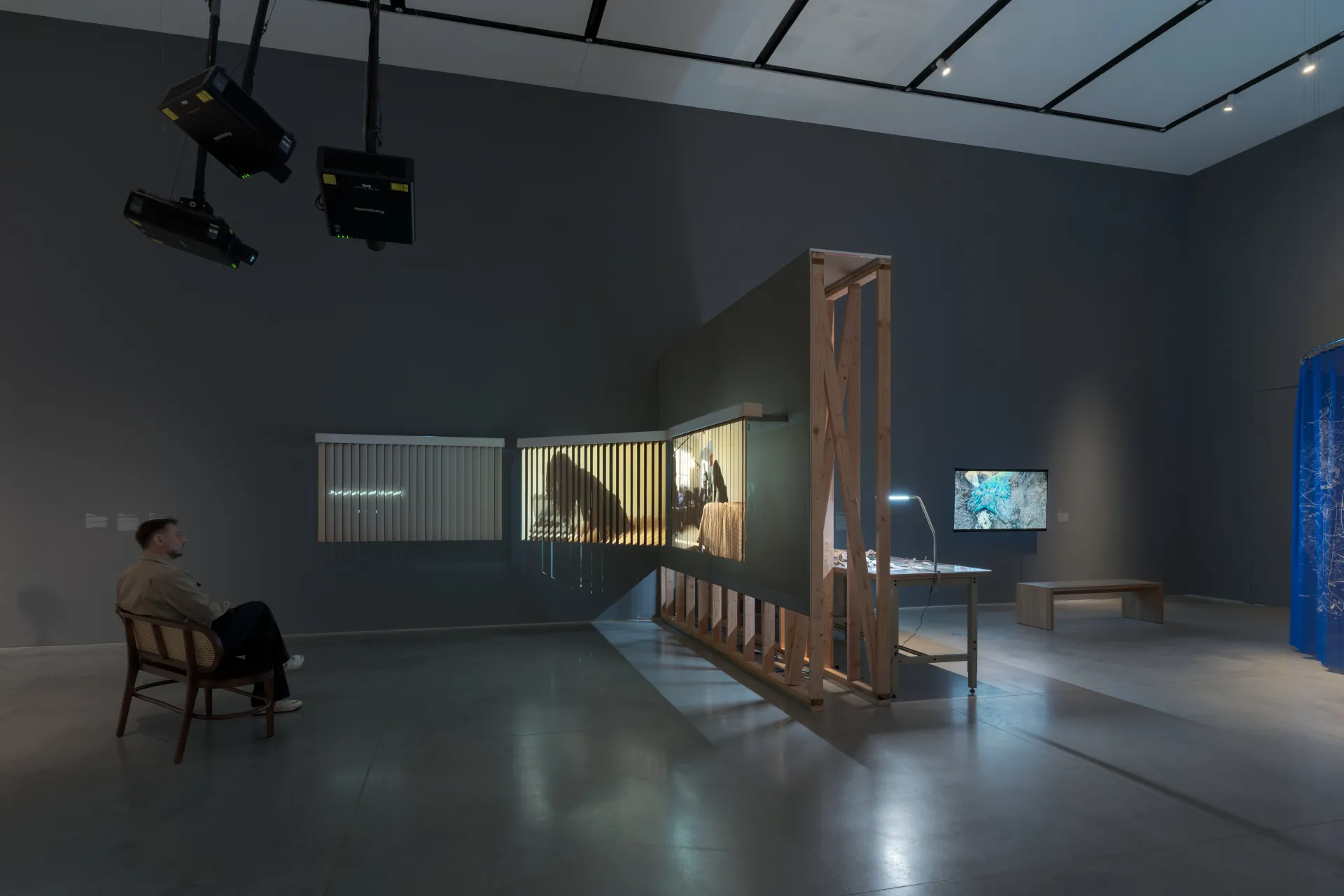 A gallery exhibition with multiple artworks including projections, a video monitor, and a table installation. A man sits on a bench watching a video work projected on blinds.