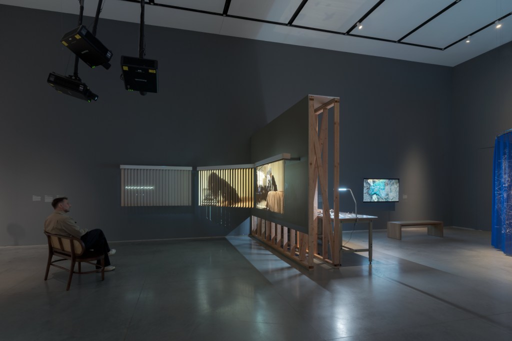 A gallery exhibition with multiple artworks including projections, a video monitor, and a table installation. A man sits on a bench watching a video work projected on blinds.