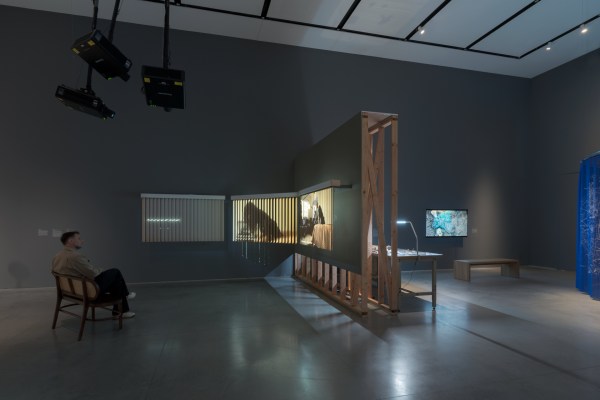 A gallery exhibition with multiple artworks including projections, a video monitor, and a table installation. A man sits on a bench watching a video work projected on blinds.