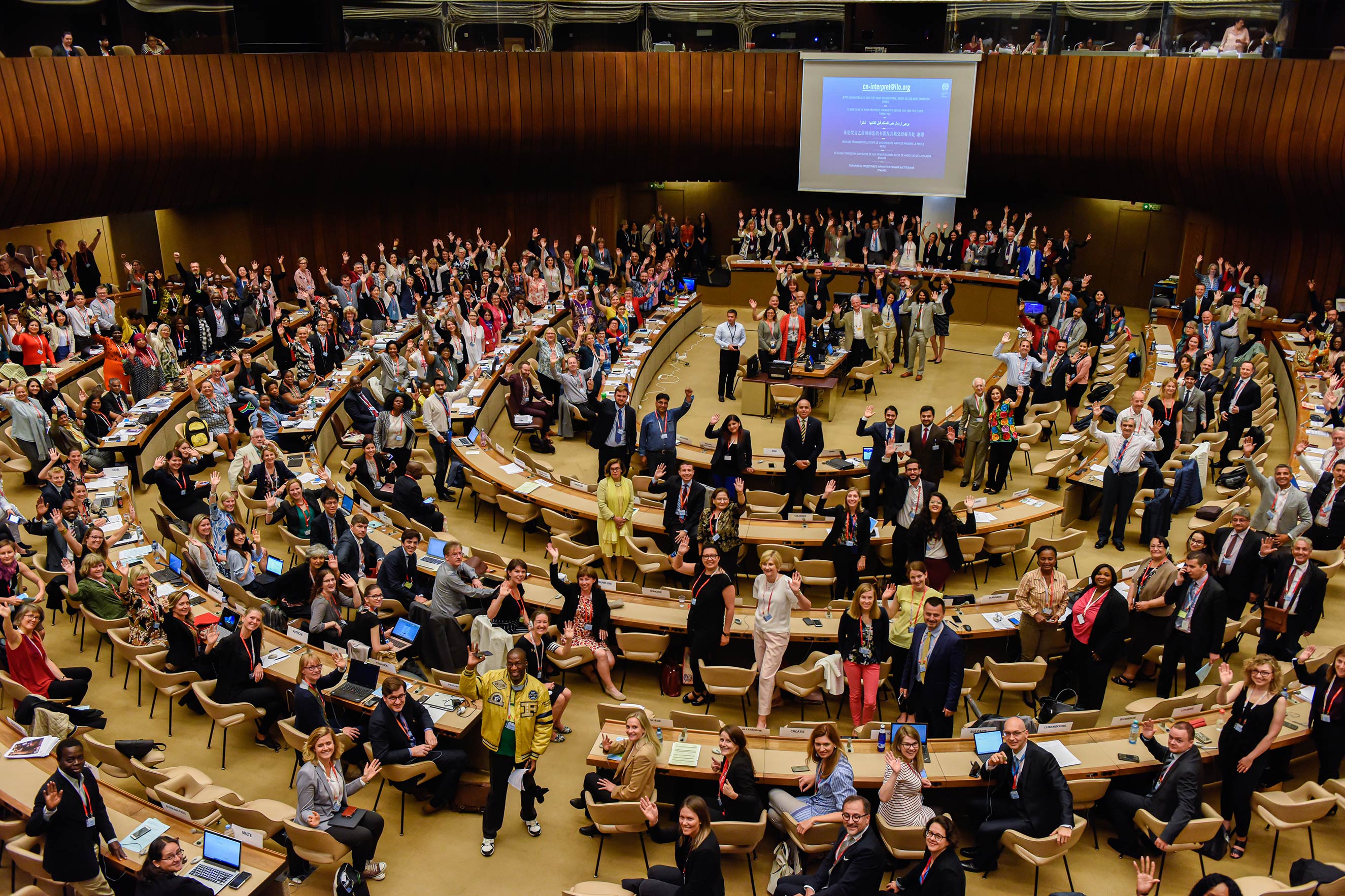 A wide shot of a large, diverse audience in a circular assembly hall with their hands raised. This action-oriented, journalistic photo captures a moment of collective participation and engagement, humanizing the foundation’s work at a global labor summit.