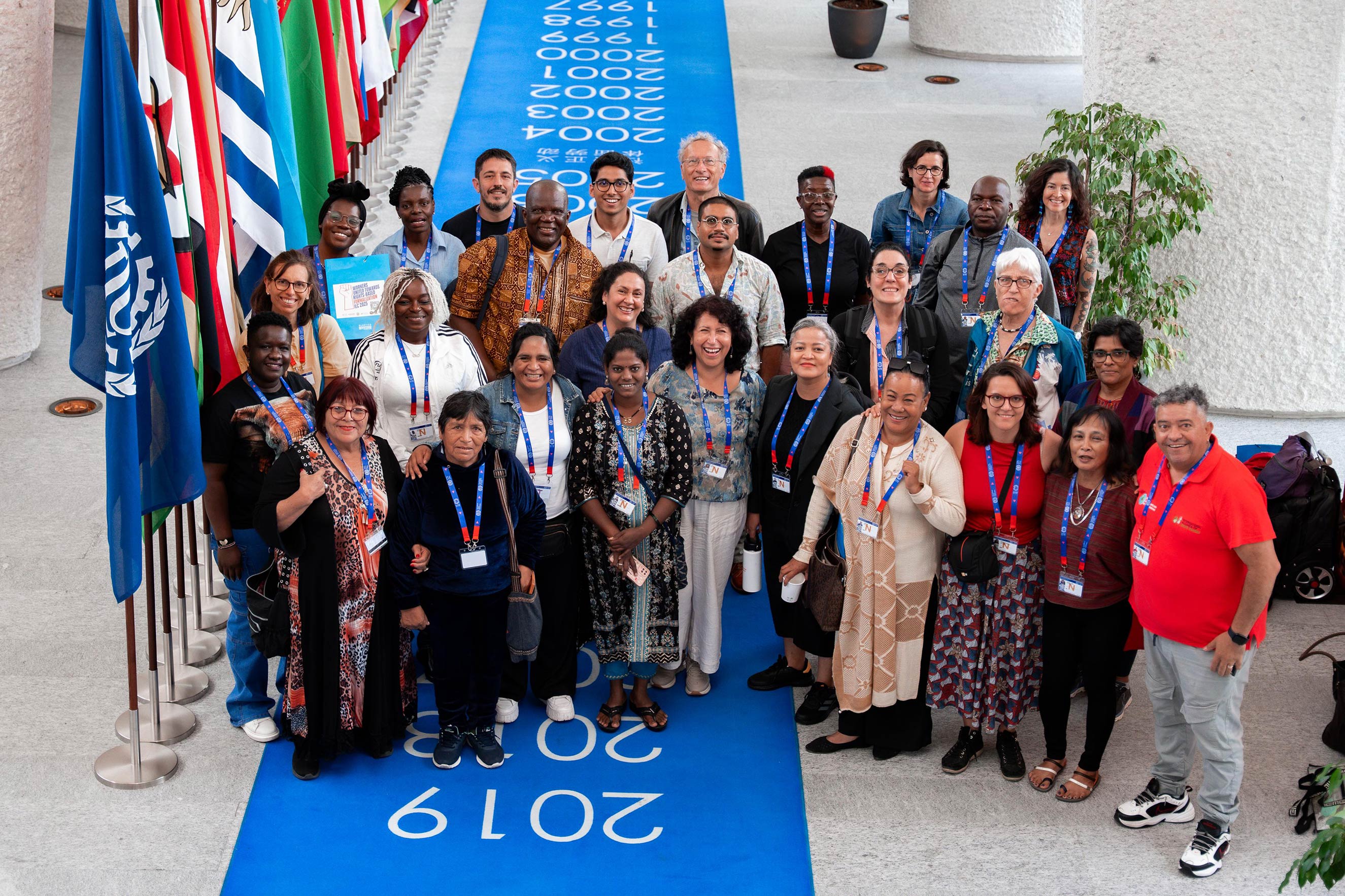 A group portrait of over 20 diverse advocates posing on a blue runner featuring chronological years. International flags line the left side. This journalistic photo humanizes the foundation’s work by capturing a moment of collective partnership at a global labor event.
