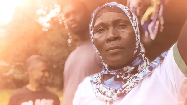 A close-up portrait of Mrs. Jalloh wearing a blue and white patterned headscarf, looking directly at the camera with a steadfast expression. Two men stand slightly out of focus in the background under warm, flared sunlight. This high-quality photo humanizes the work of legal advocates.