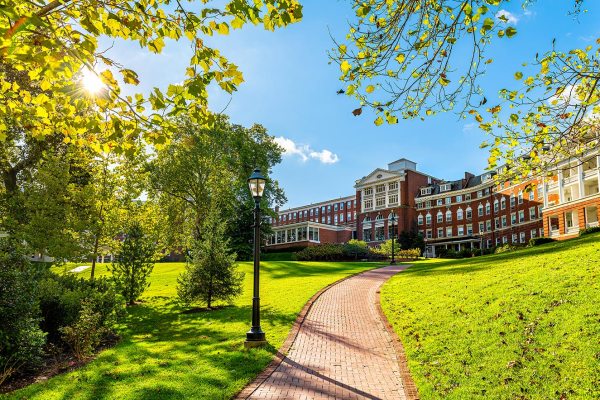 A photo of a winding brick pathway leading toward a grand, red-brick university building on a lush green campus. Sunlight filters through autumn leaves, creating a hopeful and vibrant atmosphere that enhances the story of the next generation of investment talent in Appalachia.