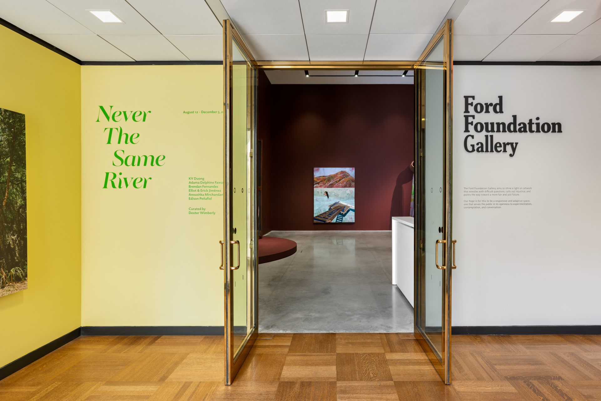 Gold-framed glass doors leading into a gallery space. To the left, a yellow wall displays green text reading 'Never The Same River' and exhibition details. To the right, a white wall has black text for 'Ford Foundation Gallery' and a description. Inside, a framed painting artwork hangs on the red wall.
