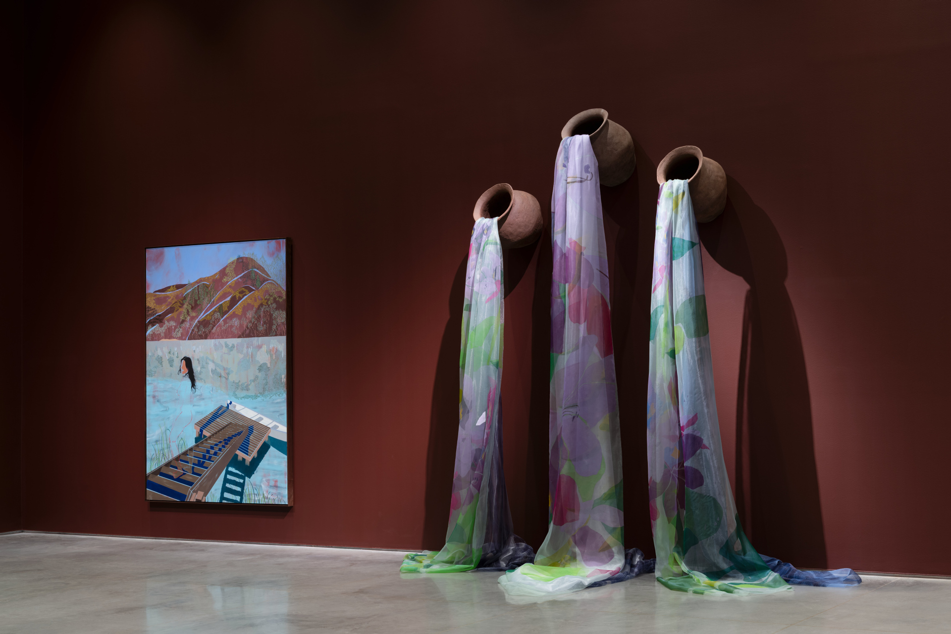A gallery wall displaying a painting of mountains above a figure in water with blue geometric shapes. To its right, three ceramic pots are mounted on the wall, each with sheer, floral-patterned fabric in shades of purple, green, and blue draped to the floor.
