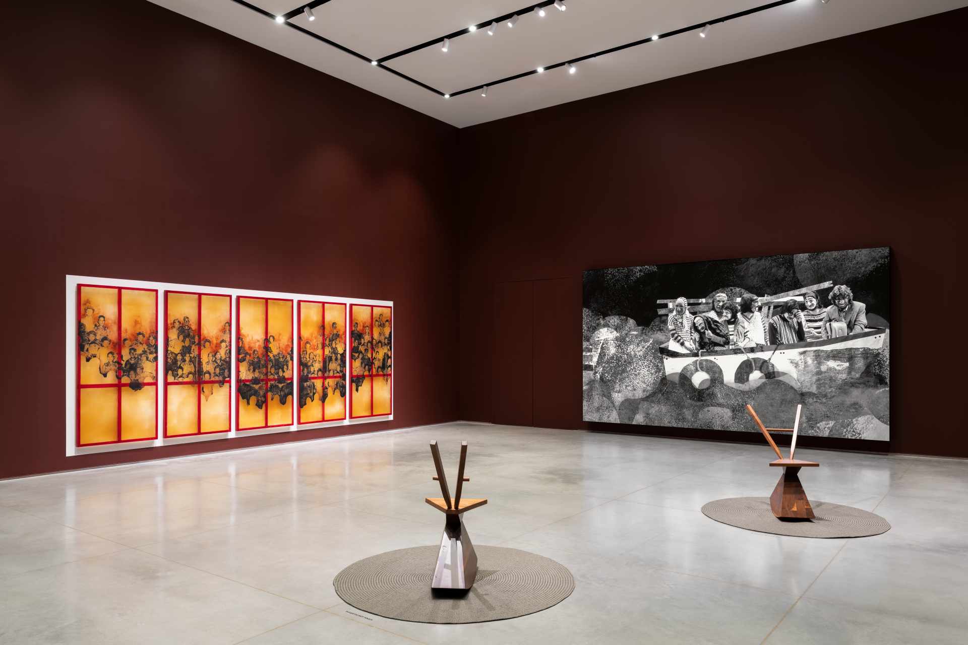 A gallery exhibition displaying multiple artworks. On the left wall is a five-panel artwork with screen-prints over a reddish-orange backgrounds . On the right wall is a large flat screen playing a video of several people on a boat. On the gallery cement floor, two geometric chair sculptures stand on circular rugs.