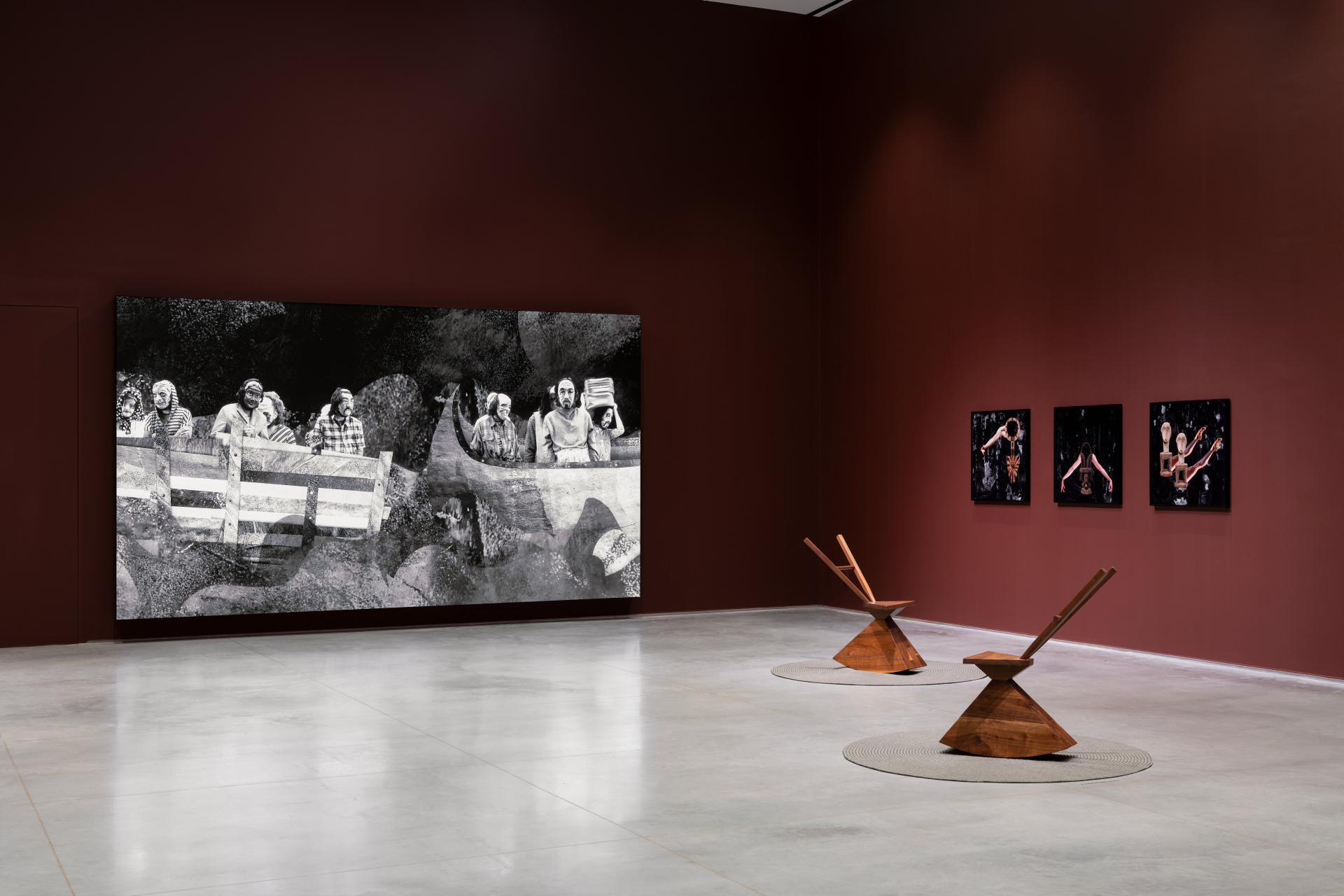 A gallery interior with various artworks. A large, horizontal screen on the left wall depicts a grey-scale video of several people traveling on a river in a wooden raft-like structure. Two abstract wooden chairs with wide bases and narrow backrests sit on circular gray rugs in the center, and three smaller, dark framed artworks hang on the right wall.