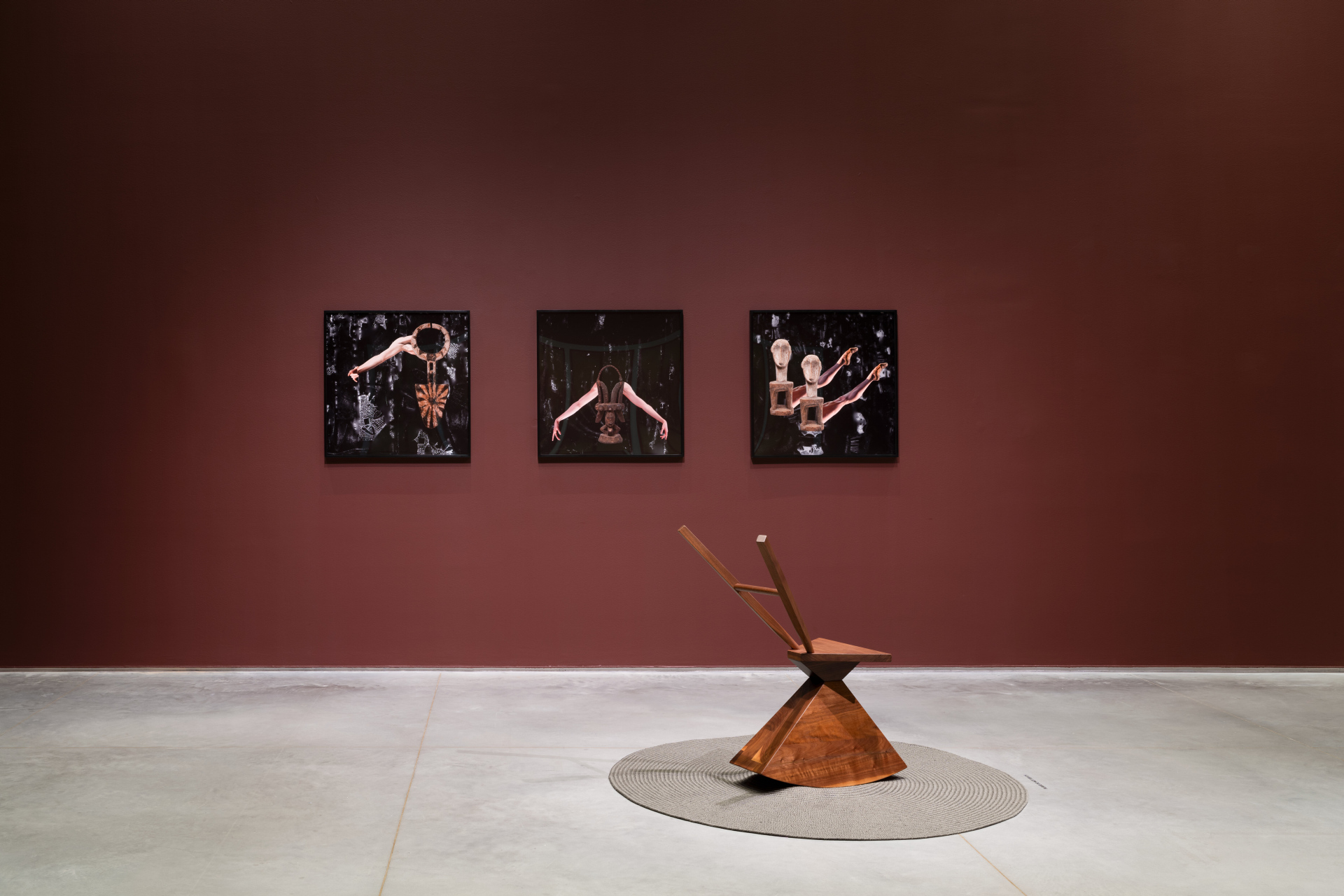 Three dark framed artworks hang on a dark red gallery wall. Each artwork features an artifact juxtaposing ballet limbs against a dark background. Below them, a wooden sculptural chair with a triangular base and an angled backrest rests on a circular gray mat.