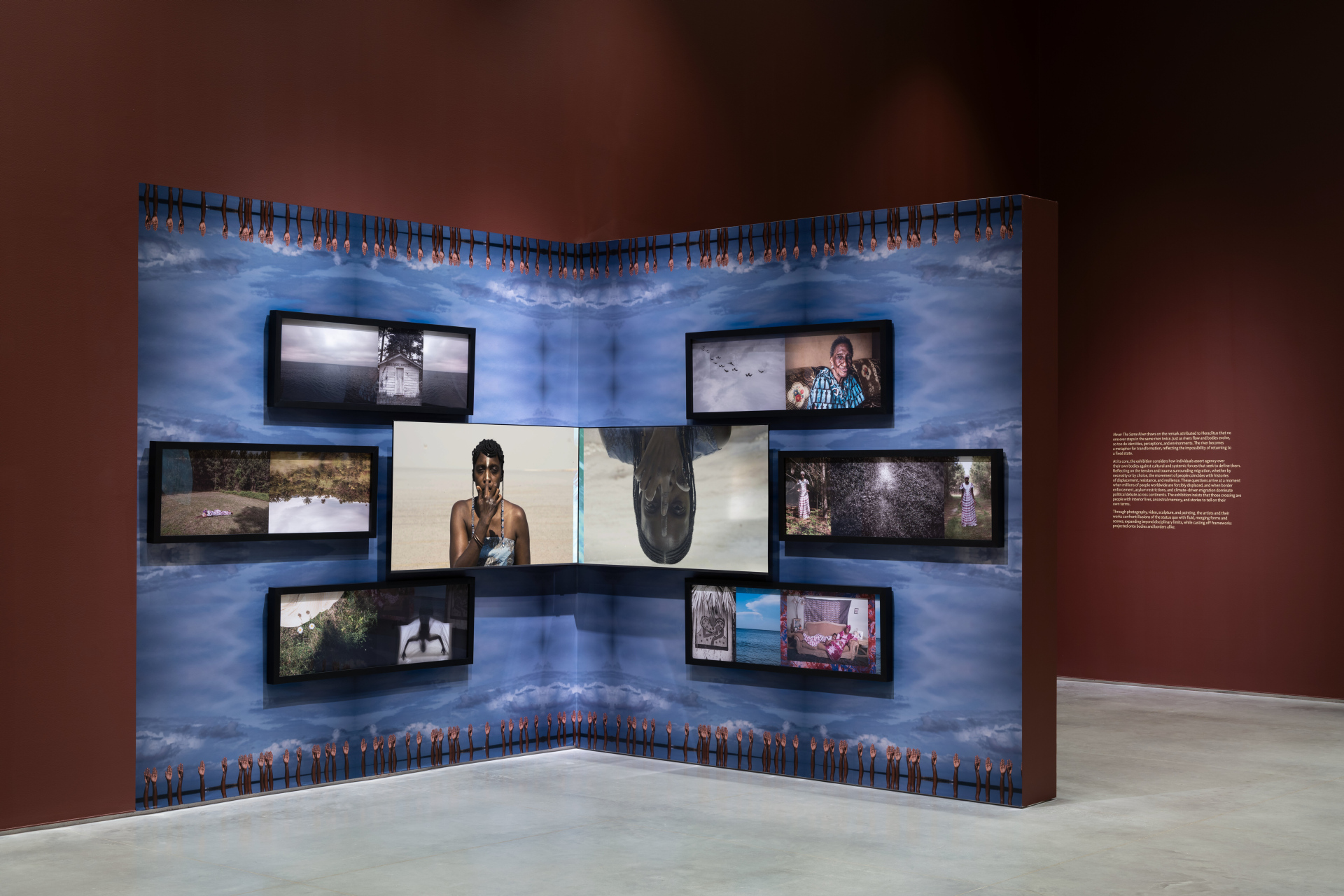 A gallery installation on a corner wall with blue and white patterned wallpaper that has a repeating border of hands extending towards the middle. There are various screens displaying images and videos of portraits, landscapes, and figures in water.