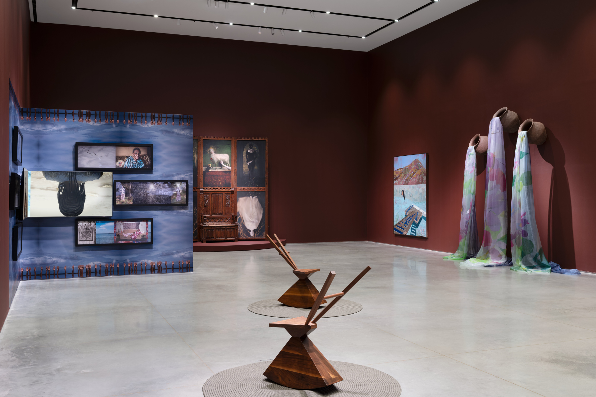 A gallery exhibition with various artworks. On the left, a blue wall installation displays multiple video screens. On the far wall, a dark wooden structure with painted canvases stands on a circular platform. In the center, two angular wooden chairs are on circular rugs, and on the right, three large vessels with flowing translucent fabrics stand beside a blue abstract painting.