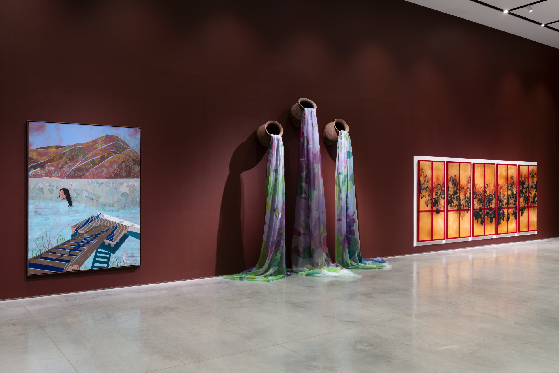 An art gallery featuring three distinct installations on a dark red wall. On the left, an abstract painting depicts a person standing beside a lake, beneath a landscape of brown hills. In the center, three ceramic pots are mounted on the wall, with sheer, colorful fabrics appearing to pour from them onto the floor. To the right, five framed panels display dark, intricate designs against a vibrant red-orange background.