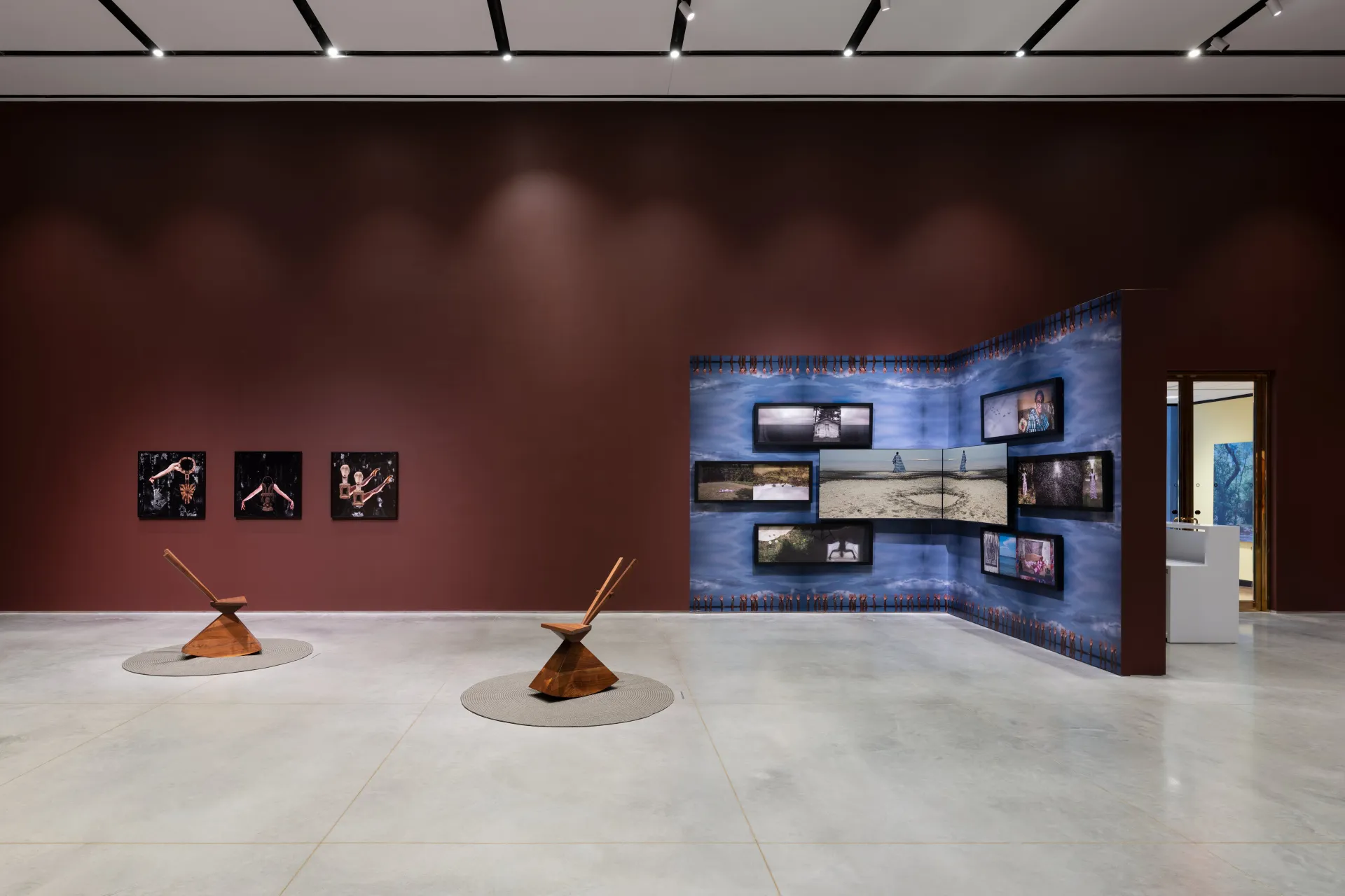 A gallery exhibition with various artworks. From left to right, there are three framed prints, two sculptural wooden chairs, a corner-wall installation with blue patterned wallpaper and various framed photographs, and an exit.