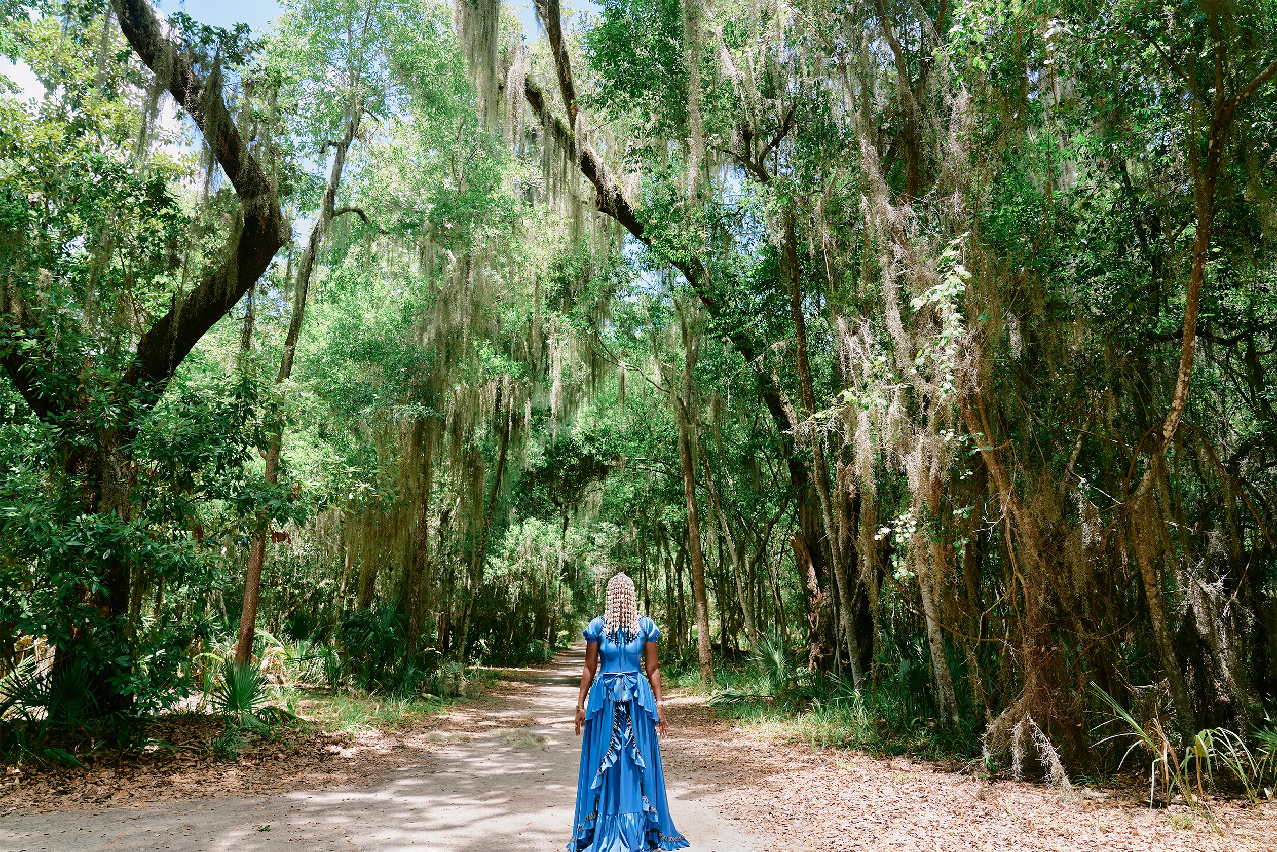 A Black woman in a long and flowing, blue dress stands on a dirt pathway surrounded by a forest of tall, mossy trees. Her beaded hair drapes over her eyes, concealing most of her face.