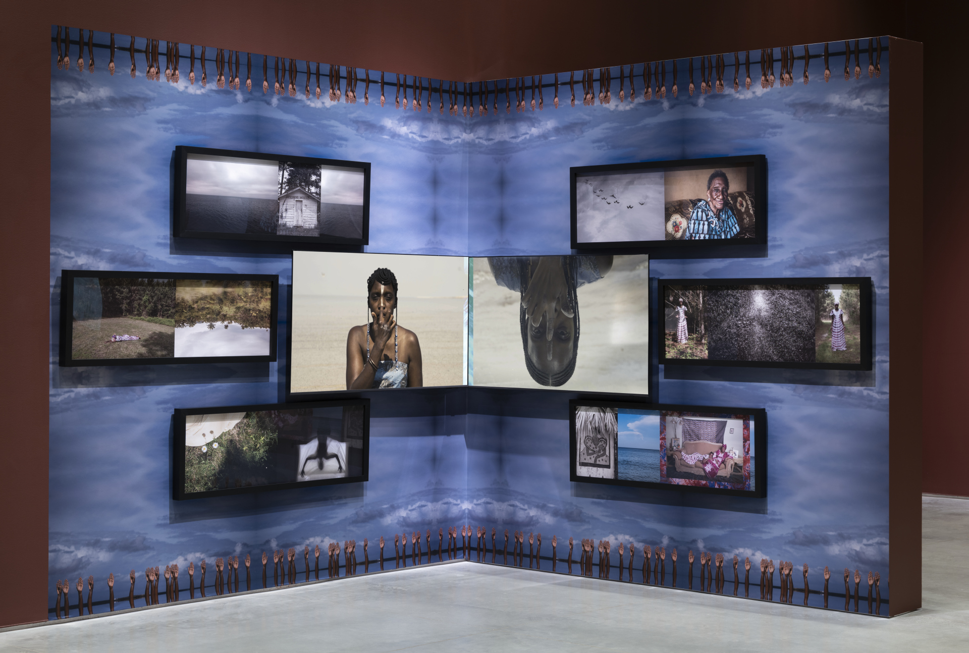 An art installation in a gallery corner featuring two walls covered in a blue, cloud-like patterned wallpaper. On the top and bottom border of the wallpaper are a row of hands extending towards the middle of the installation. Multiple framed images and tv monitors are displayed, and mounted so that each wall mirrors the other.