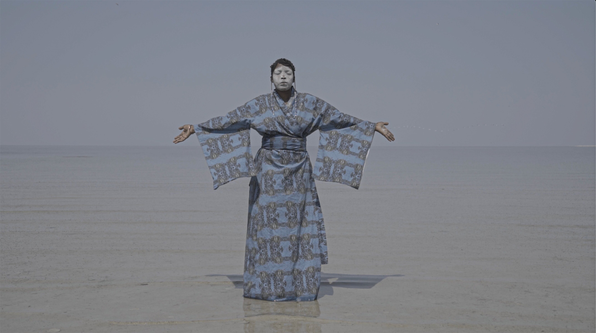 A Black woman wearing a light blue patterned robe with wide sleeves and a cinched waist stands with arms outstretched and palms open. White paint covers her face as she stands on a beach under a pale sky.