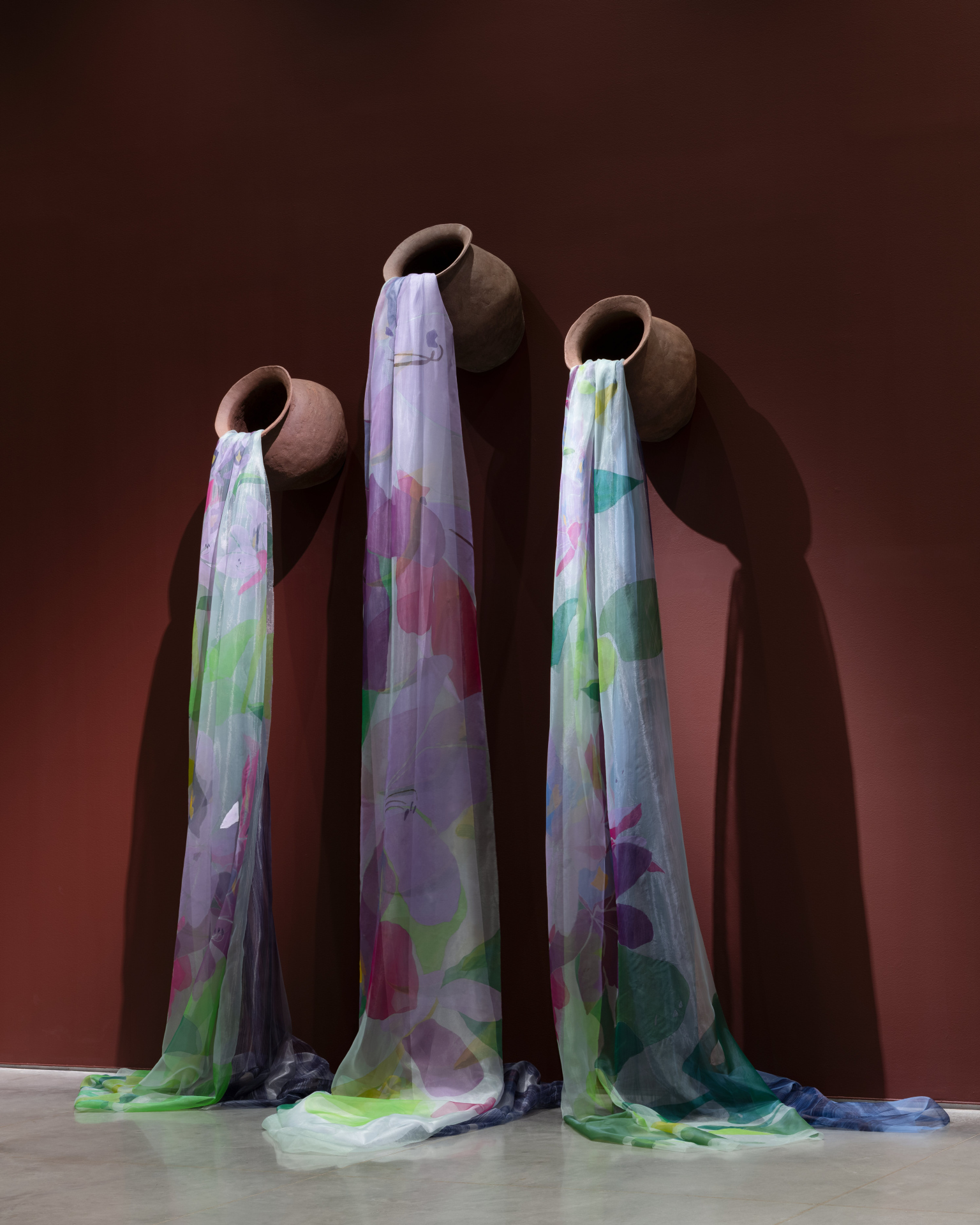 Three unglazed ceramic pots mounted on a burgundy wall, each spilling a long, sheer fabric to the floor. The fabrics are painted with purple, green, and blue floral designs.