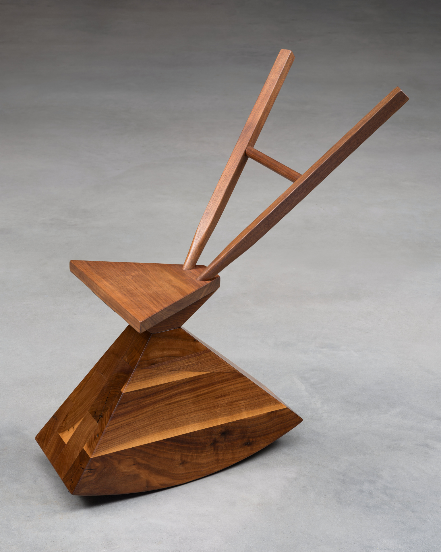 A dark wood chair, composed of geometric shapes. A wide, angled block forms the base, topped by a triangular seat. Two long, angled wooden bars extend upwards from the seat, joined by a horizontal bar.