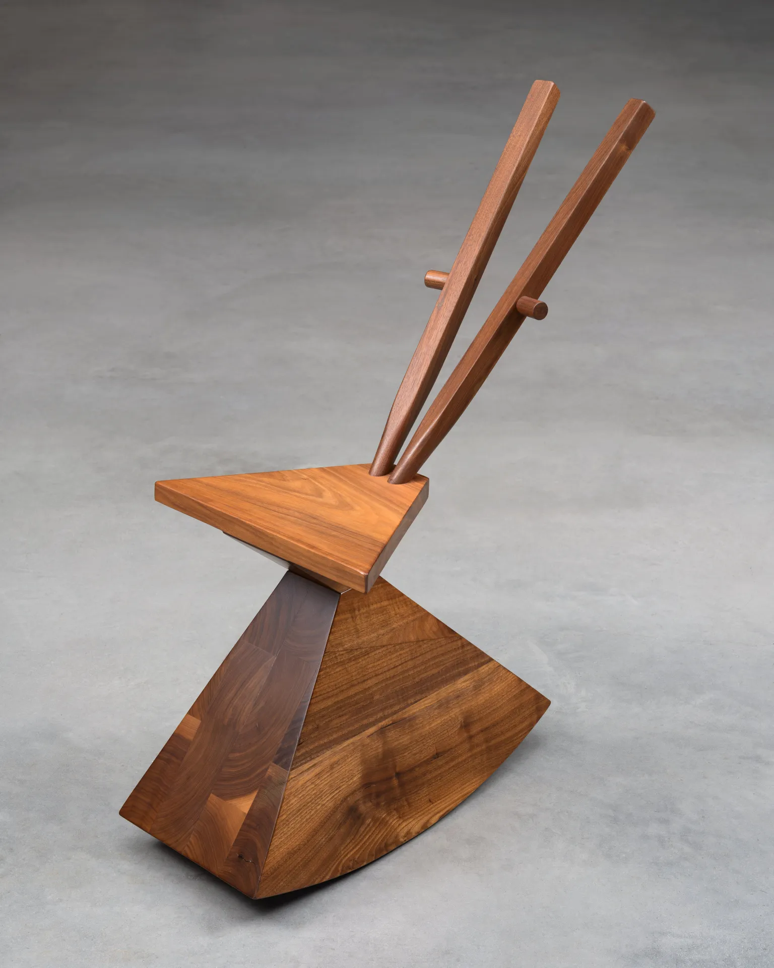 A sculptural wooden chair with a geometric design, facing towards the bottom left corner. It features a faceted, inverted pyramid base, a lighter triangular seat, and two slender wooden rods extending upwards from the back, each with a small peg.