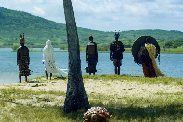 Five individuals in distinct, symbolic traditional attire stand on a grassy shoreline looking out over a wide river or lake. A tree trunk dominates the foreground, framed by rolling green hills in a scene from a Ford Foundation JustFilms documentary.