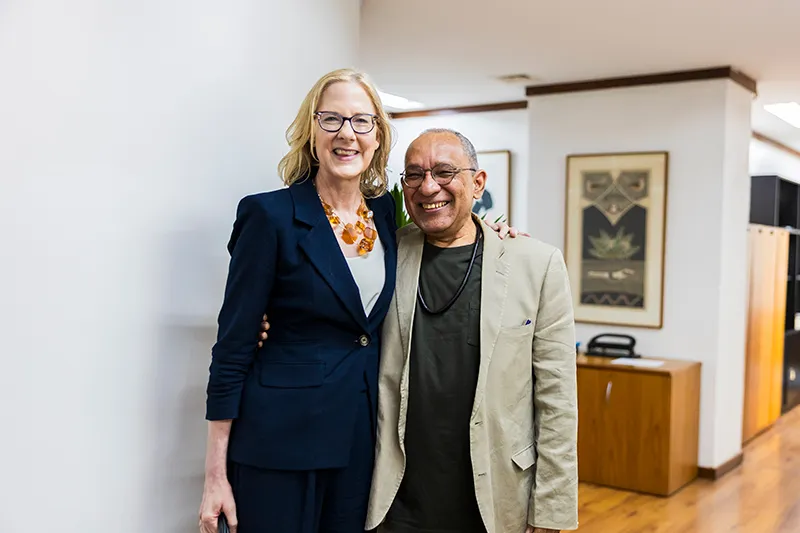 Heather Gerken poses with Átila Roque. Heather smiles wearing glasses, a blue blazer, and an amber necklace. Átila is wearing a beige blazer, glasses, and a black beaded necklace. The setting is professional.