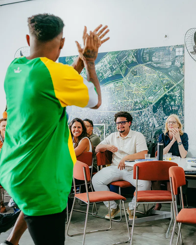 A man in a green and yellow shirt claps in the foreground. Sitting in the background, Heather Gerken and others watch and smile in front of a large map.