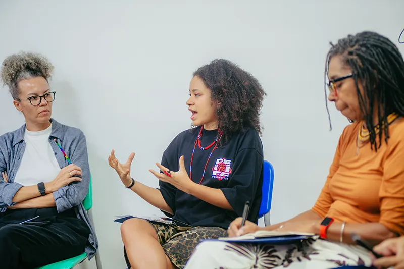 Three individuals engage in a discussion while seated in chairs. They each hold notebooks, and one is speaking with expressive hand gestures. The group appears focused and attentive.
