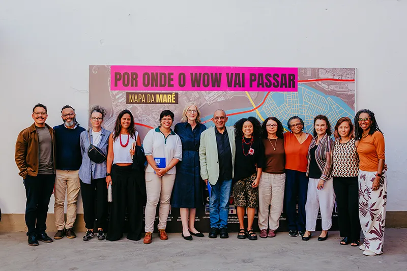 Heather stands at the center of a group of people in front of a large map labeled "Mapa da Maré" and "Por Onde o Wow Vai Passar" in pink. They are arranged in a line, smiling at the camera. The background is a plain white wall.
