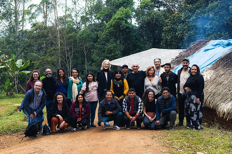 A group of people poses in front of a rustic building with a thatched roof, surrounded by lush greenery.