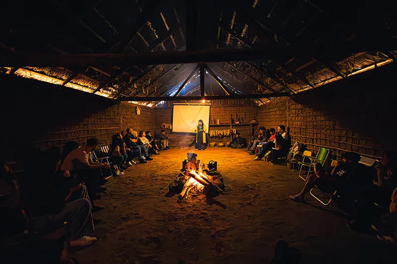 A person stands in front of a screen inside a dimly lit thatched building with a campfire in the center. People are seated in a circle around the fire, attentively watching the screen. The interior has a rustic and communal atmosphere.