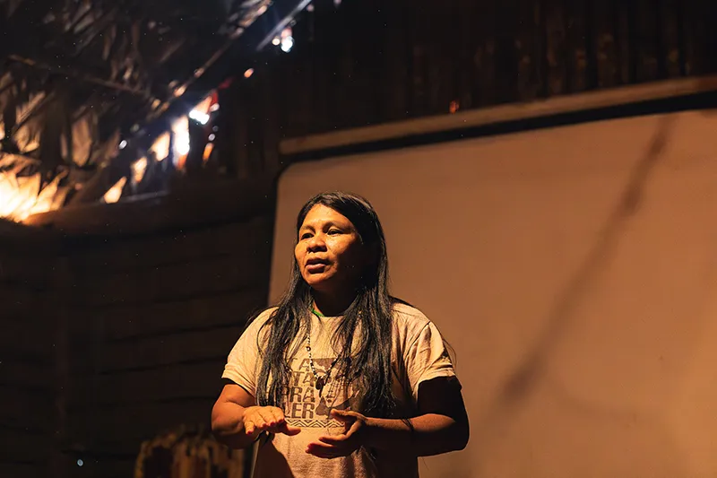 A person stands indoors, speaking passionately. She’s wearing a t-shirt and gesturing with her hands. The setting has wooden walls and dim lighting, suggesting an intimate atmosphere.