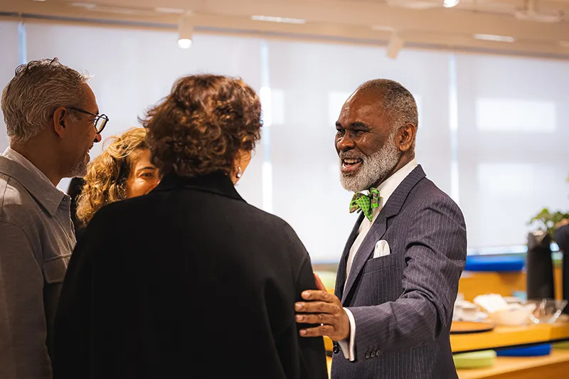 A smiling Black man with a grey beard, wearing a suit and green bow tie, talks warmly to a group of people at an indoor gathering.