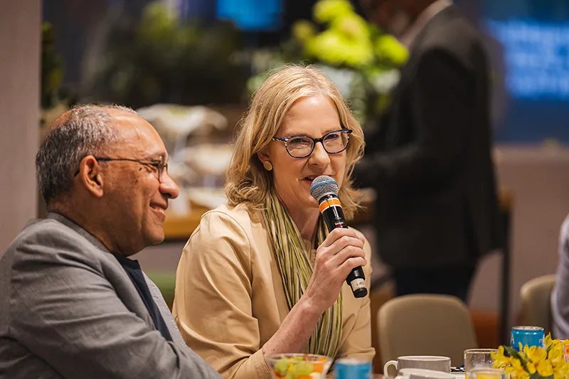 Heather Gerken speaks into a microphone while sitting next to a smiling Átila Roque, in a grey suit, during an indoor event.