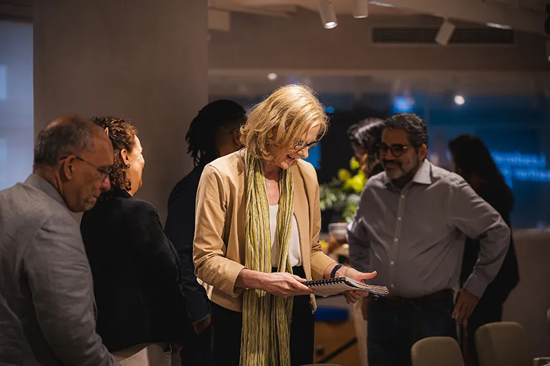 Heather holds a bound booklet and smiles while other individuals look on. She is wearing a tan blazer and green scarf. The setting is in a large room with floral arrangements.