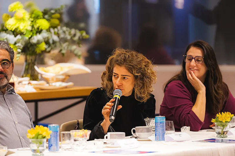 A person speaking into a microphone at a conference table with others seated nearby. There are floral arrangements and beverages on the table.