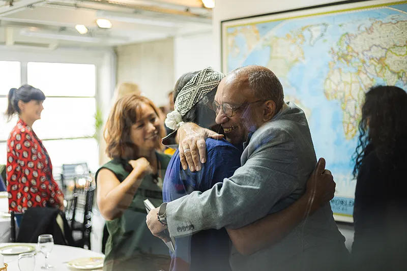 Átila Roque, wearing a grey jacket, smiles warmly as he shares a big hug with someone in front of a world map during a gathering.