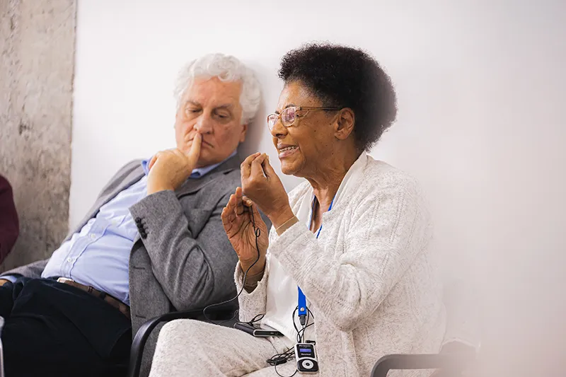 An older Black woman speaks and gestures expressively while a white-haired man in a grey blazer listens thoughtfully beside her at an event.