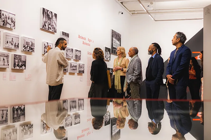 A group of people stands in an art gallery at the Zumvi Photography Exhibition, observing black-and-white photographs displayed on the walls. The room is well-lit, with reflections visible on a glossy surface in the foreground.