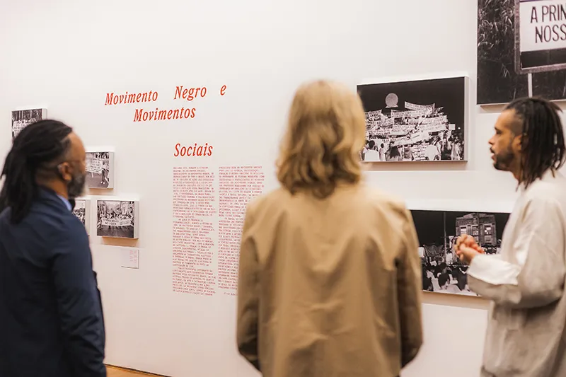 Three individuals are observing the Zumvi Photography Exhibition featuring black-and-white photographs. The text on the wall reads "Movimento Negro e Movimentos Sociais" in red. The setting appears to be a gallery or museum.