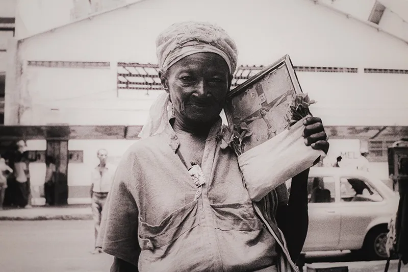A black-and-white photo from the Zumvi Photography Exhibition. In it a person is seen holding a framed photograph on a street. The background features a building and a partially visible vehicle, possibly a car.