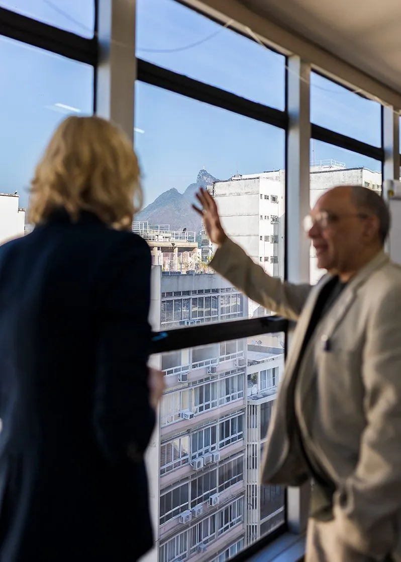 Heather and Átila stand in front of a window with a view of residential buildings and mountains and blue sky in the background. Átila raises his hand to point outside.