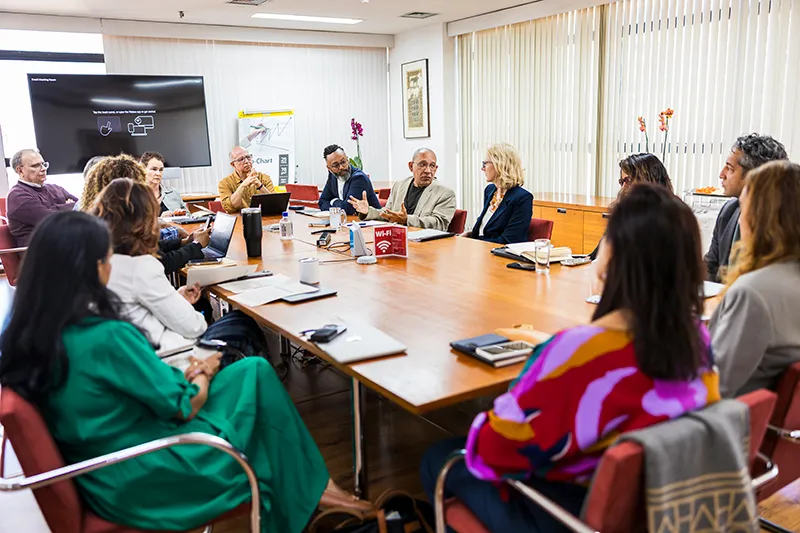 At a Ford staff meeting where individuals are seated together engaged in a discussion. They wear various colorful, professional outfits. The setting is a meeting room with a large wooden table.