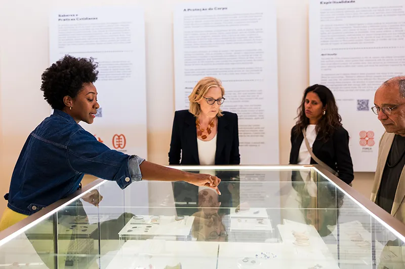 Heather Gerken and Átila Roque are standing with two other individuals in front of a glass exhibition at the Museum of Afro-Brazilian History and Culture. One individual points to an artifact at the center of the exhibition as others look on.