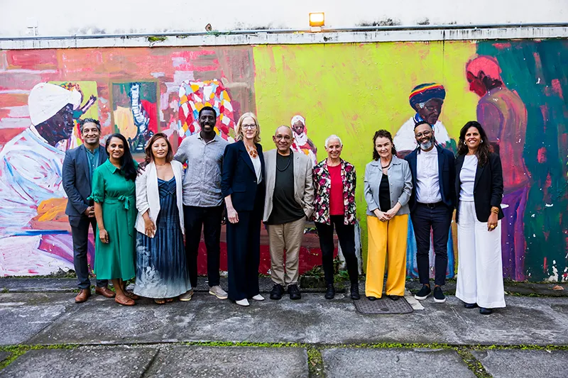 Group photo featuring Heather and Átila alongside other smiling individuals in business casual clothing in front of a mural. The mural depicts several figures with dark skin and head coverings who are standing in front of colorful patterned backgrounds of vibrant yellow, pink, and green.