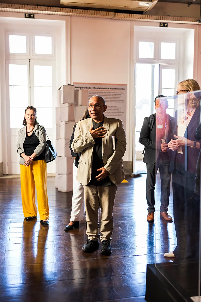 A group of people stand attentively indoors, where Átila is gesturing in conversation. They are in a well-lit room with wooden floors and large windows. Museum display cases and informational panels can be seen in the background.