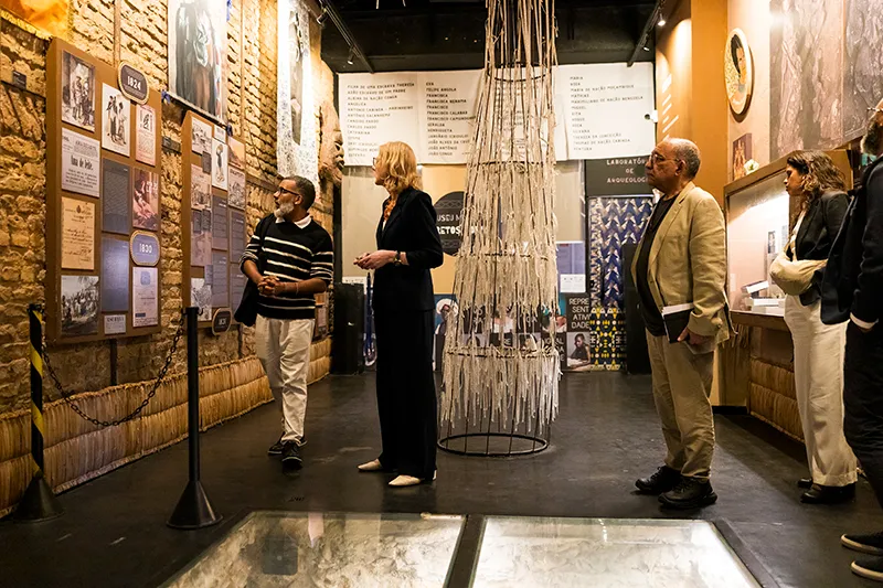 Heather stands among a group of people observing exhibits at the Instituto Pretos Novos. The room has warm lighting and exhibits on the walls with photographs and text. There is a tall, cylindrical art installation in the center of the room. Exhibits feature historical information and images.
