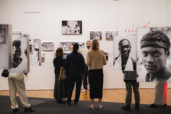 A group of people views a black-and-white photography exhibition on Afro-Brazilian culture and Candomblé traditions in Brazil. A tour guide gestures toward framed prints on the wall while large, hanging portraits flank the foreground.