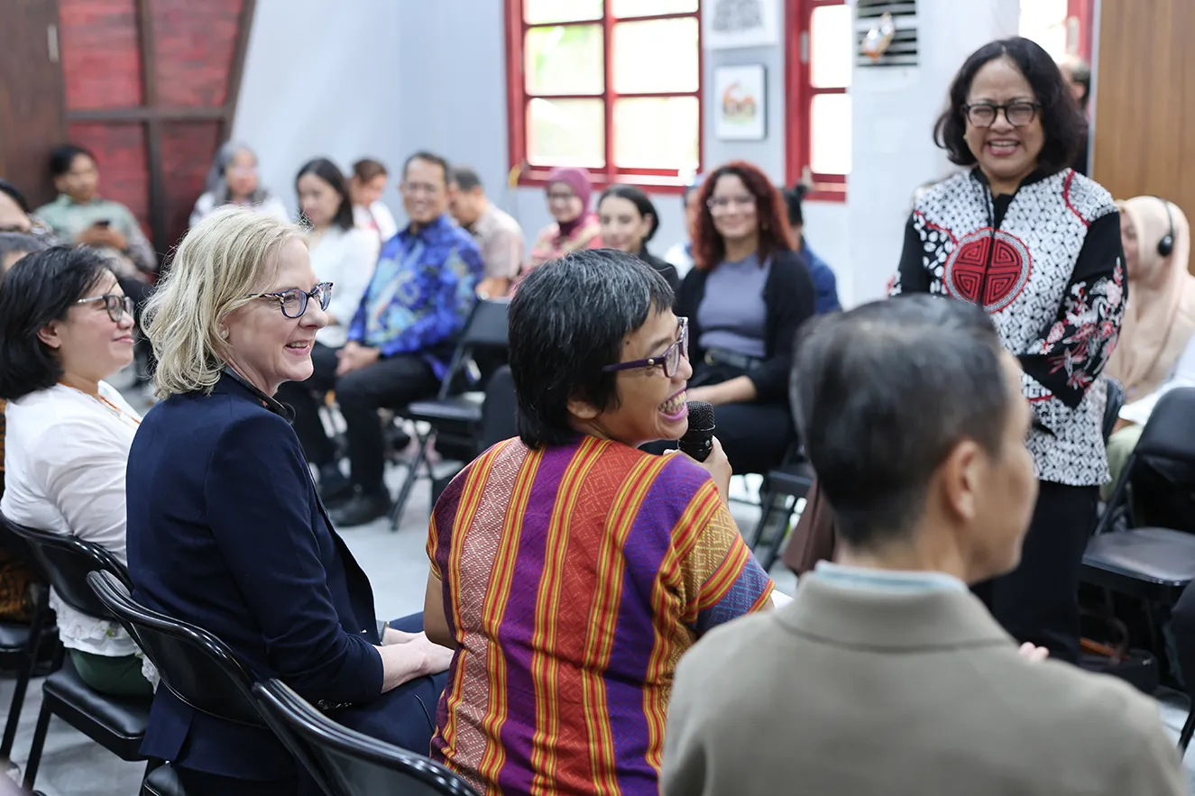 Women speaking into a microphone during an indoor regional event. Other attendees listen and smile in the background, creating an engaging, journalistic scene that humanizes the foundation's global community work in Jakarta, Indonesia.