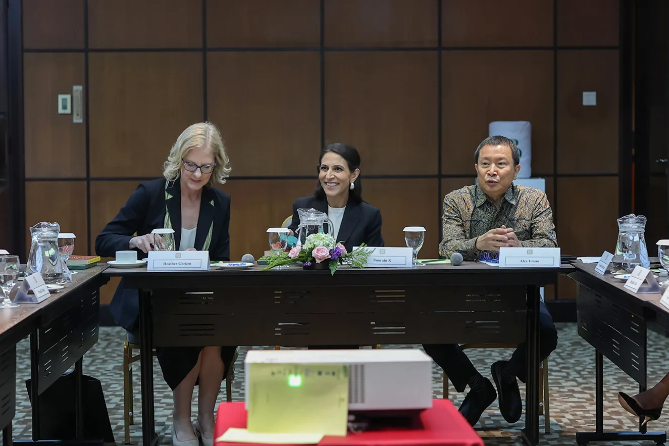 Left to right, Heather Gerken, Noorain Khan, and 
Alexander Irwan seated at a dark conference table during a regional meeting in Jakarta, Indonesia. A man in a batik shirt speaks while two colleagues smile alongside him. This journalistic image highlights institutional collaboration and humanizes our global team.