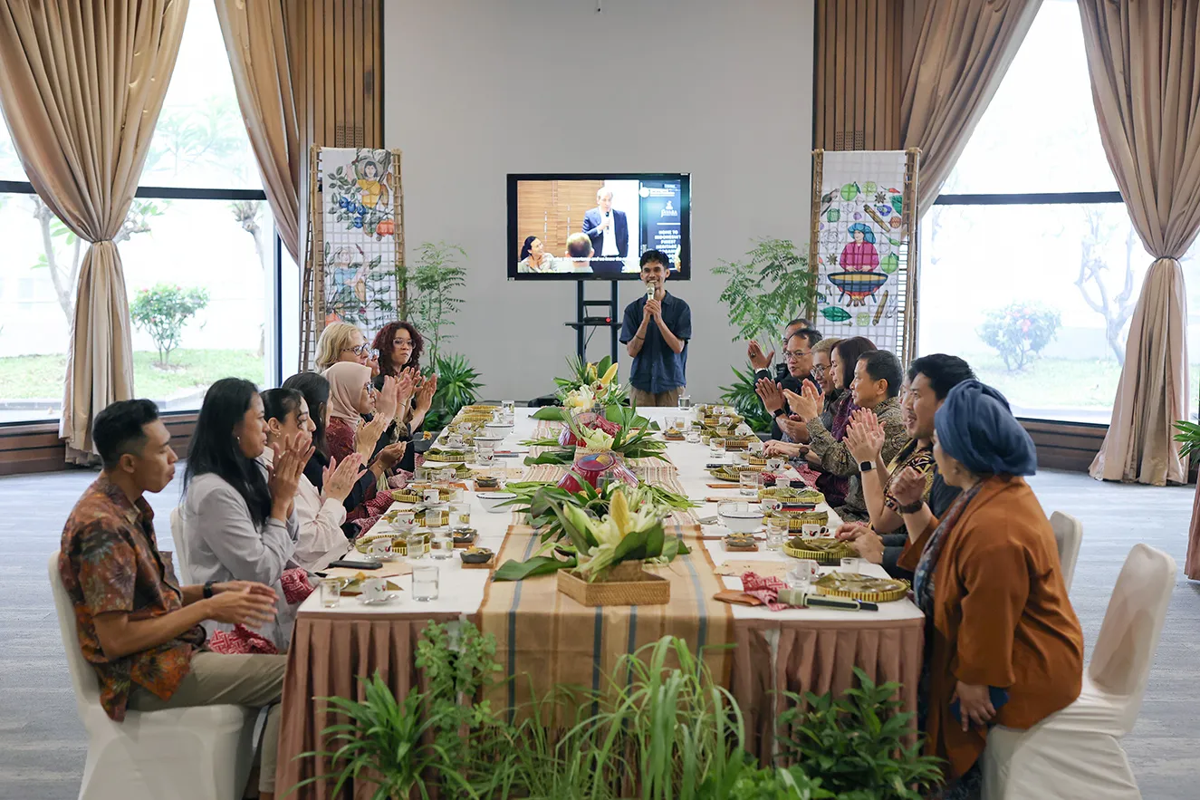 Professional leaders and staff members seated along a long, decorated banquet table in a bright room. Attendees applaud a young man speaking into a microphone at the head of the table, capturing a moment of community, gratitude, and collaboration.